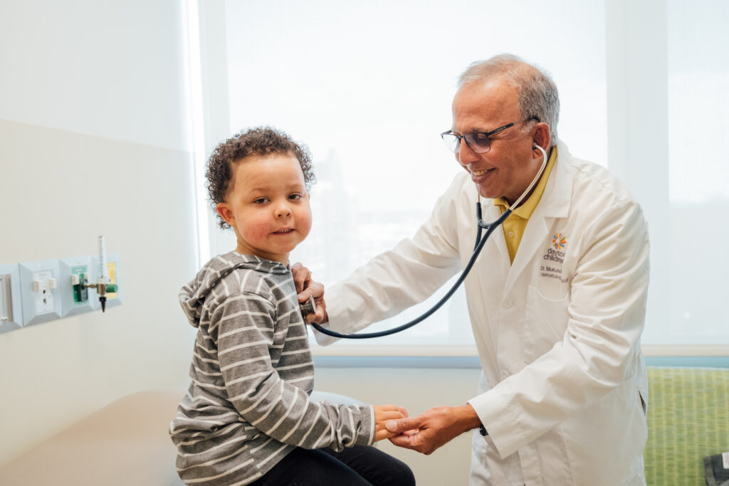 A smiling older male pediatrician listens to a young boy's heartbeat during a checkup at Dayton Children's Hospital