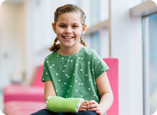 A young girl with a green cast smiles while sitting in a bright hospital lounge.