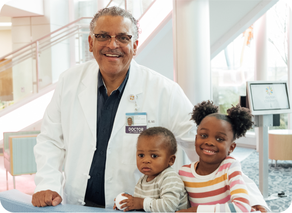 A smiling doctor stands with two young children in a hospital lobby, representing community engagement and support in pediatric care.