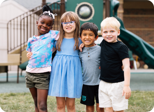 Four young children are standing in a playground outdoors with their arms around each other, smiling at the camera.