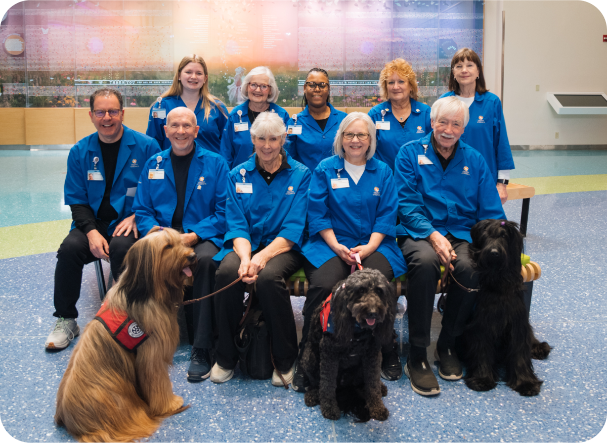 A group of volunteers, wearing their blue volunteer jackets and their volunteer dogs, sitting together smiling.