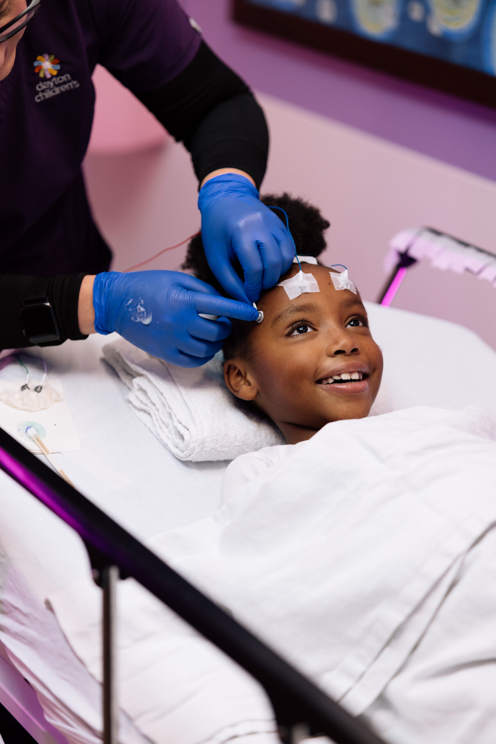 A child with a joyful expression lies on a hospital bed while a medical professional in blue gloves attaches electrodes to their head.