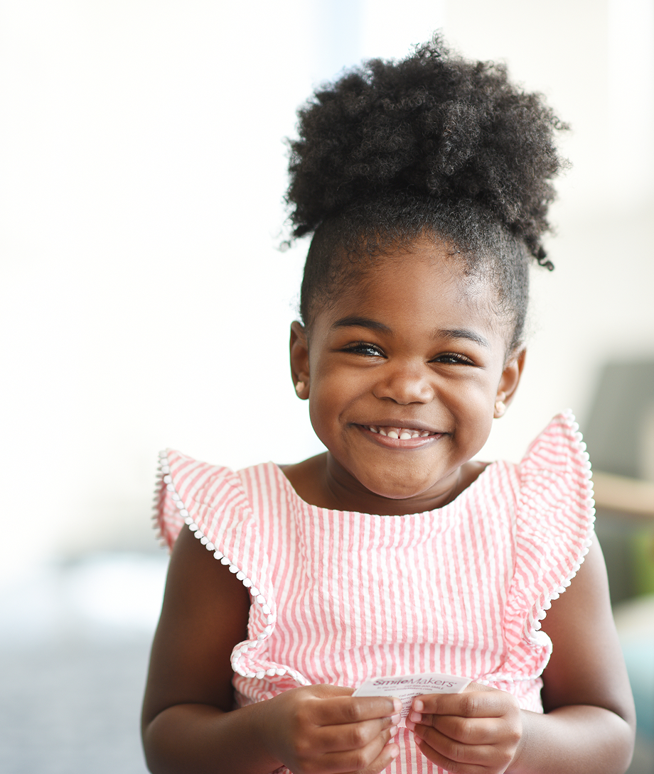 A smiling young girl in a pink top sits in a bright clinic space, representing a happy patient at Dayton Children’s.