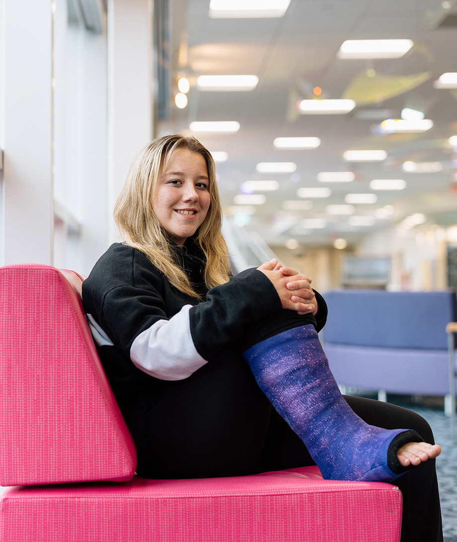 young patient wearing a purple cast on her right leg smiles while sitting on the clinic’s lobby pink couch 