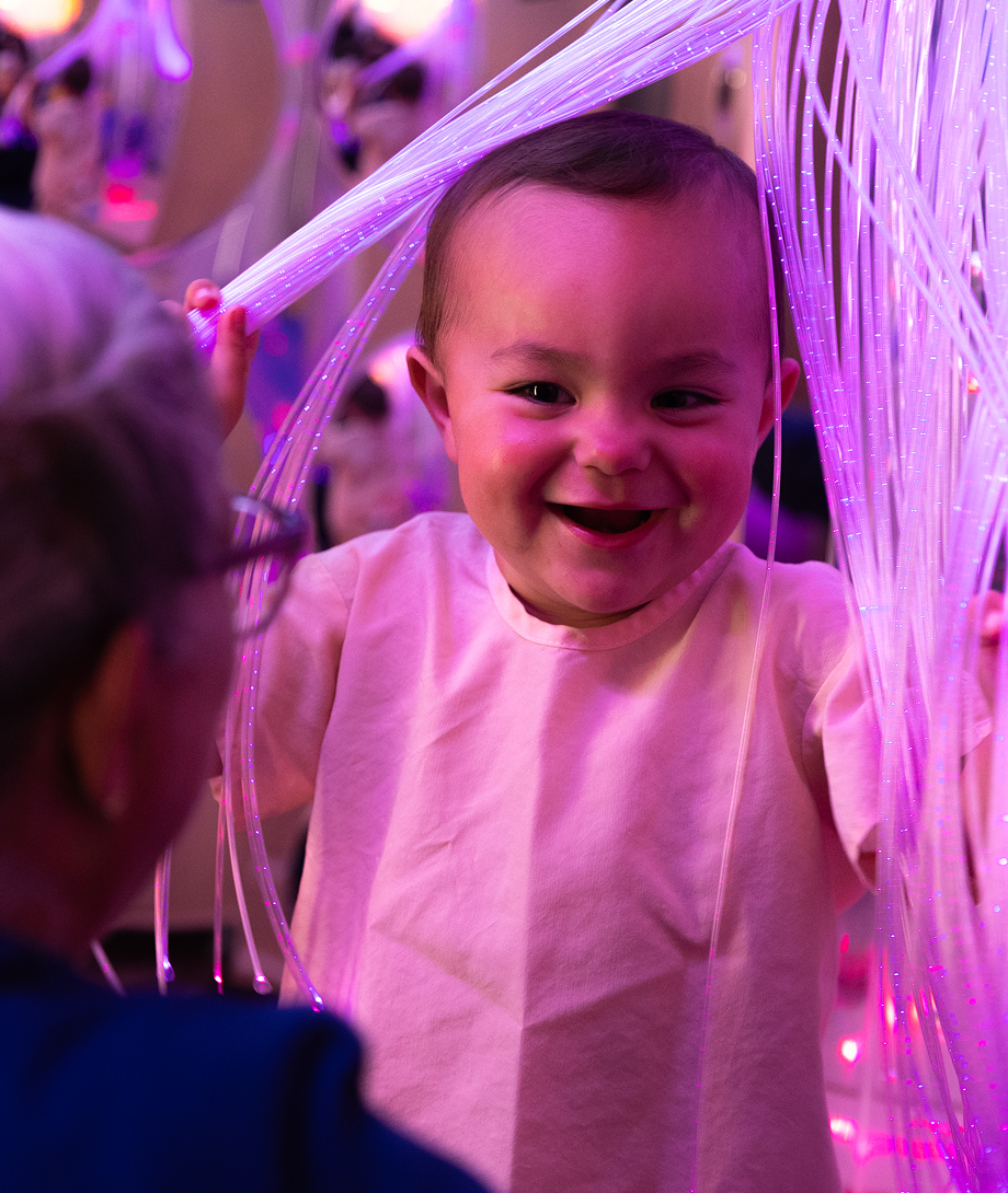 Smiling toddler playing with glowing fiber optics in a sensory room.