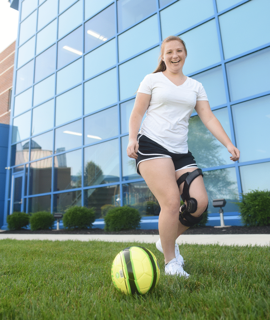 Girl kicking a neon soccer ball in grass with a brace on after her knee injury treatment
