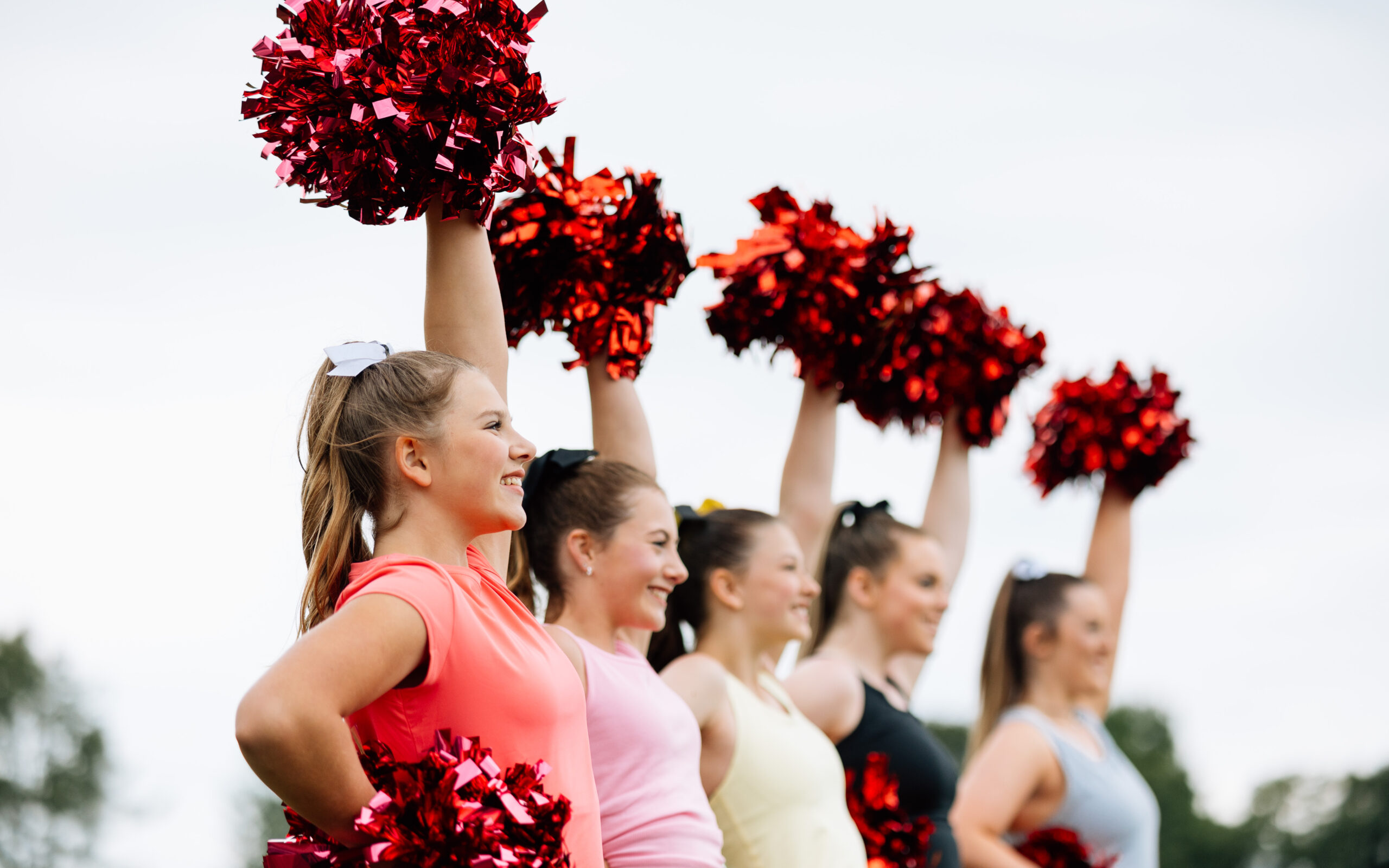 group of five female caucasian cheerleaders in a line with red pom poms, one arm up and one arm at hip