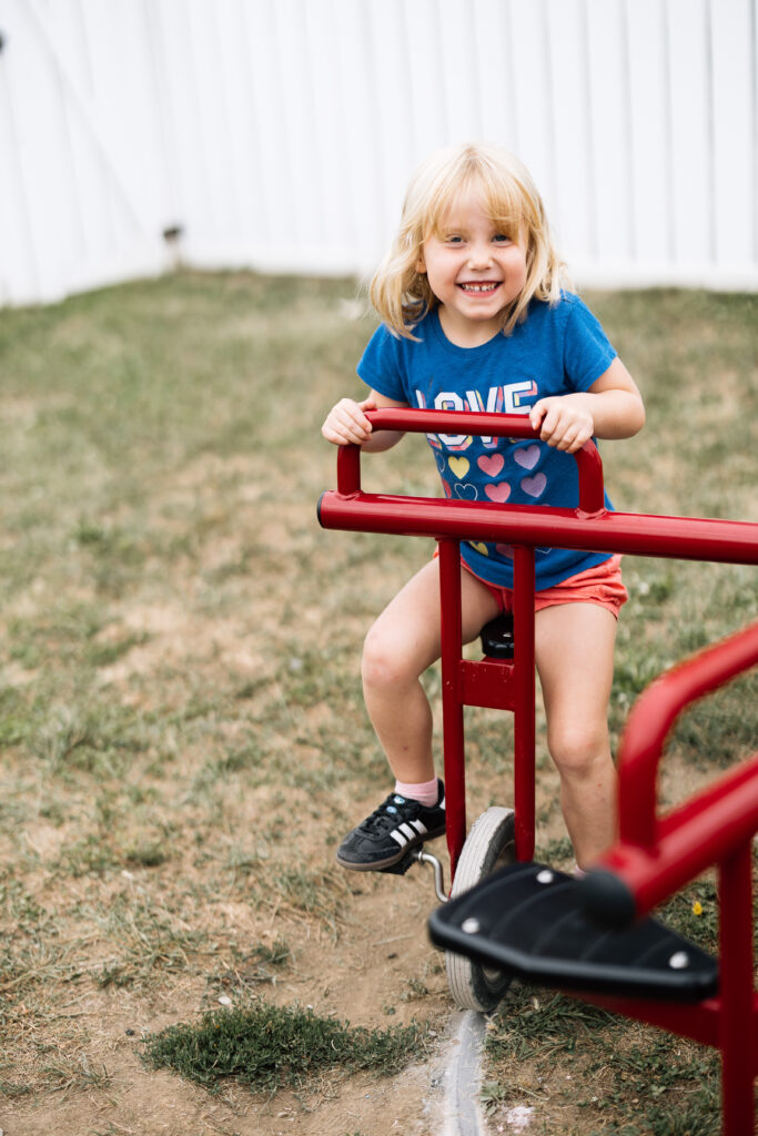 A cheerful blonde girl in a blue "LOVE" t-shirt and red shorts rides a red outdoor toy on grass.

