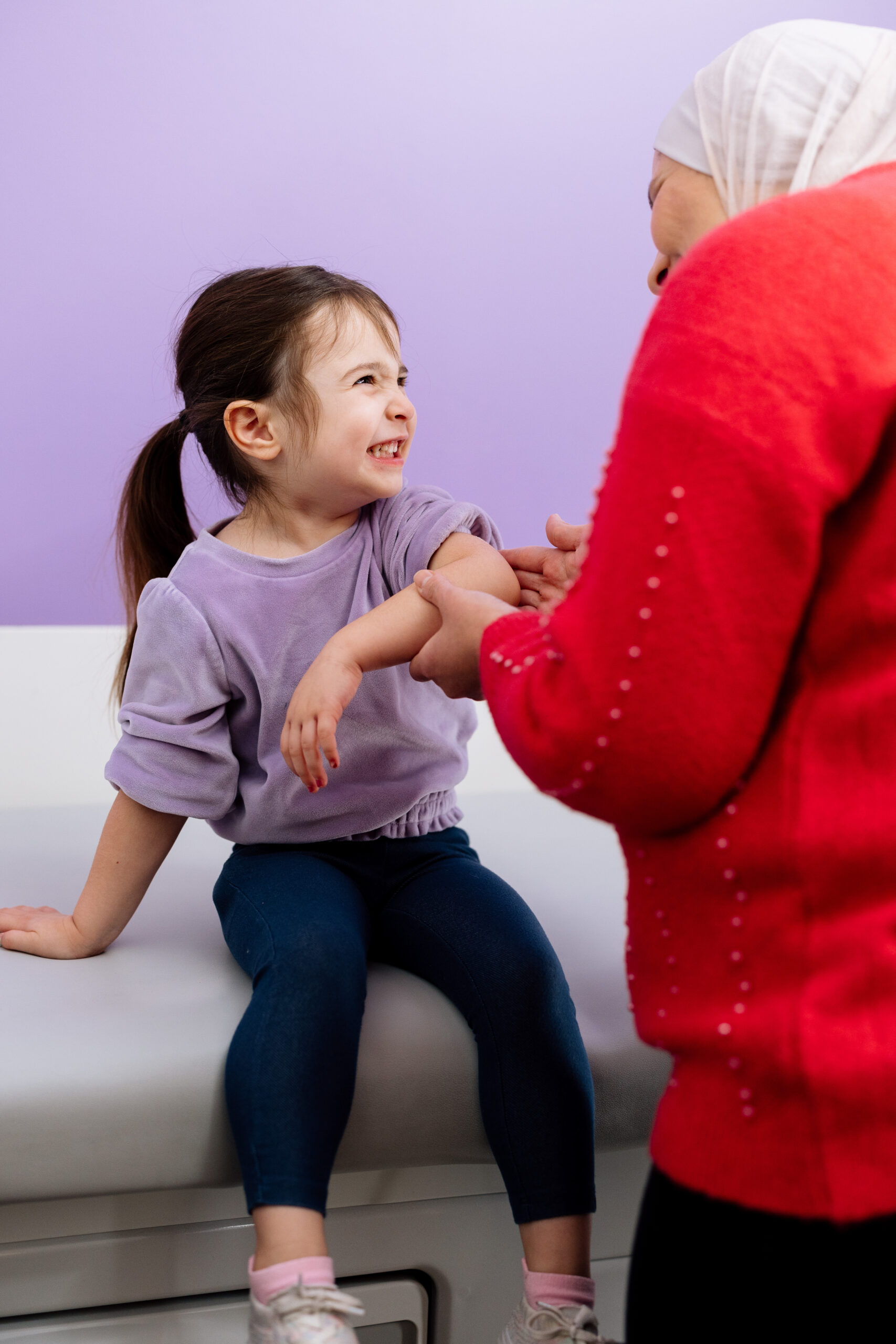 A medical professional, seen from the back, is examining a little girl's arm. The girl is smiling and laughing.