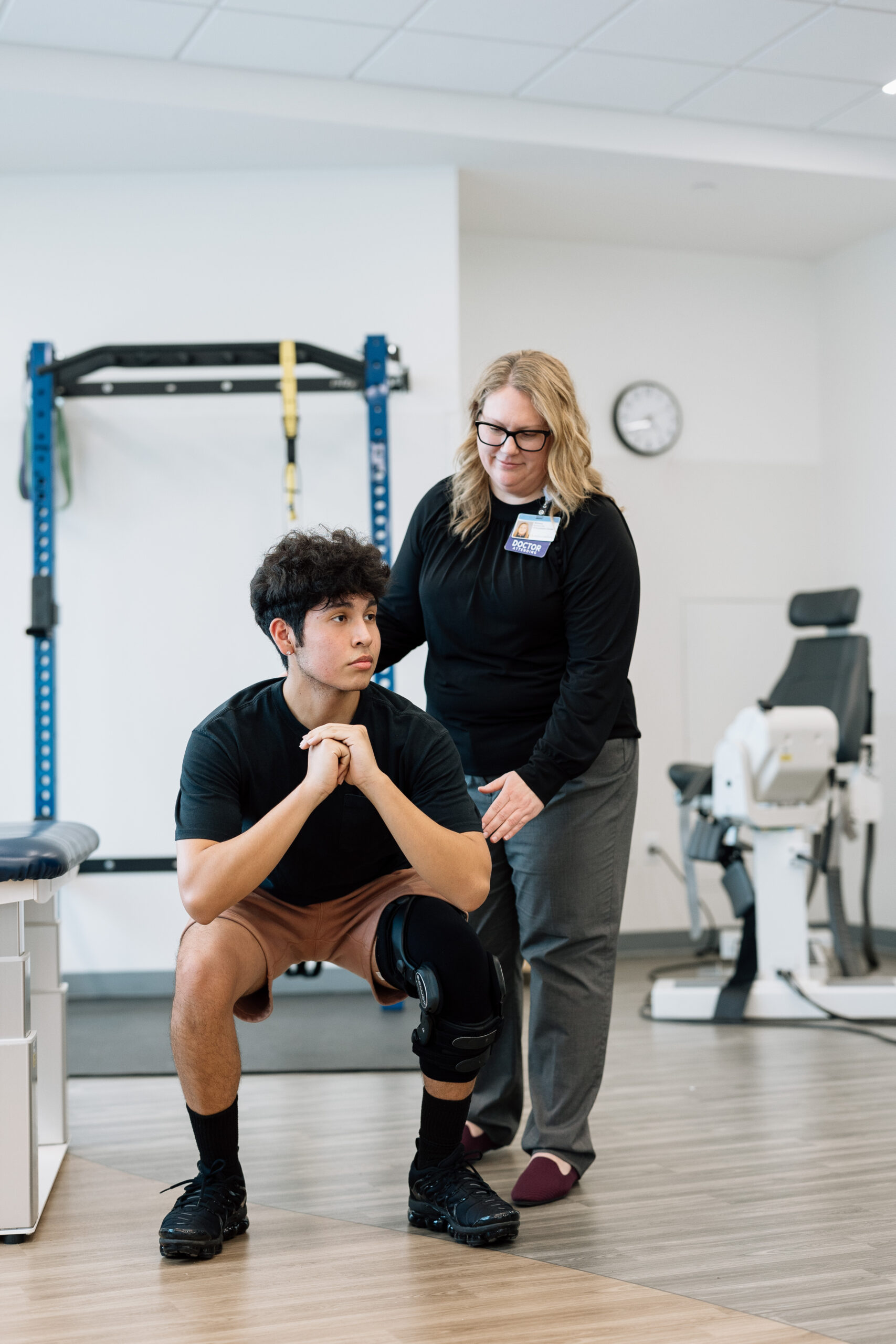 A physical therapist assists a male patient with knee injuries as he performs a squat. He is wearing a knee brace on his right leg.