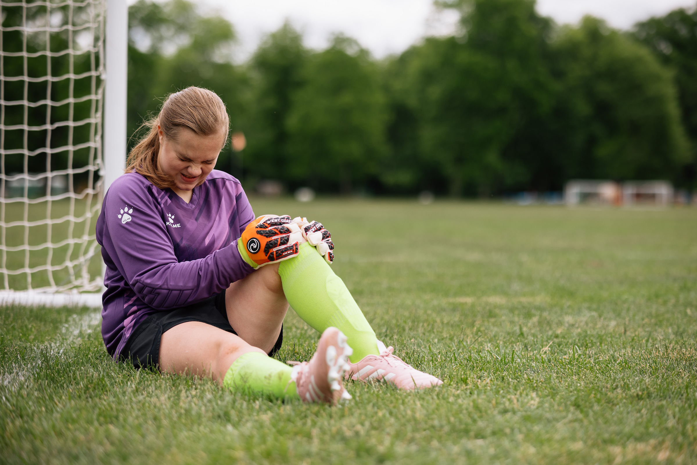 A female soccer player wearing a purple jersey and goalie gloves is sitting on a field, grimacing in pain as she holds her knee