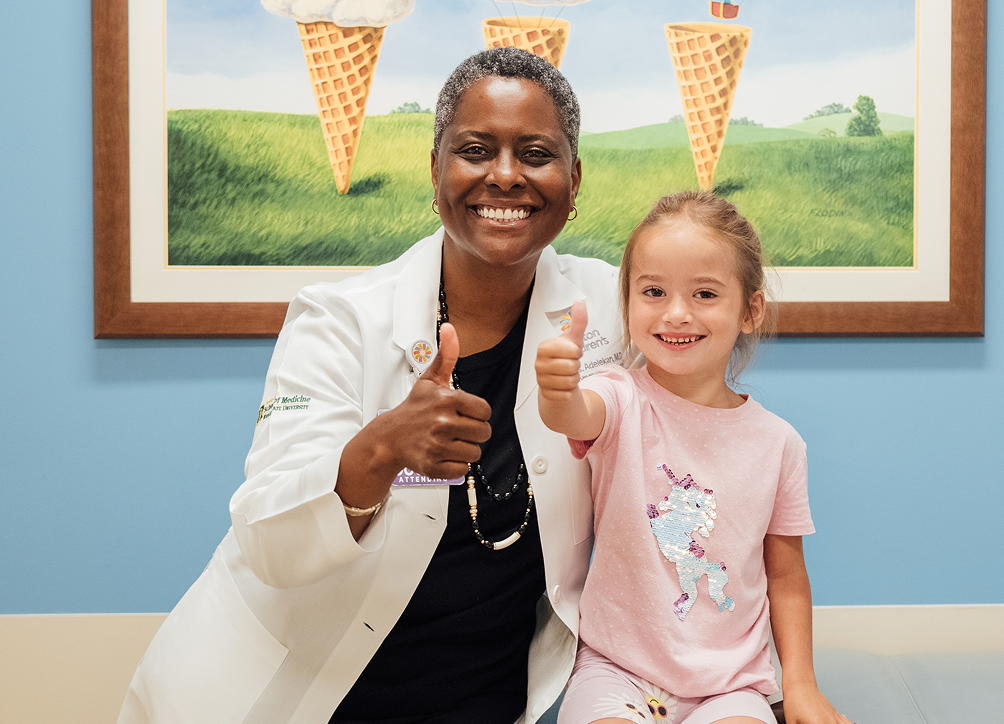 A smiling female doctor and a young girl both give thumbs up to the camera, with a whimsical ice cream cone painting behind them.