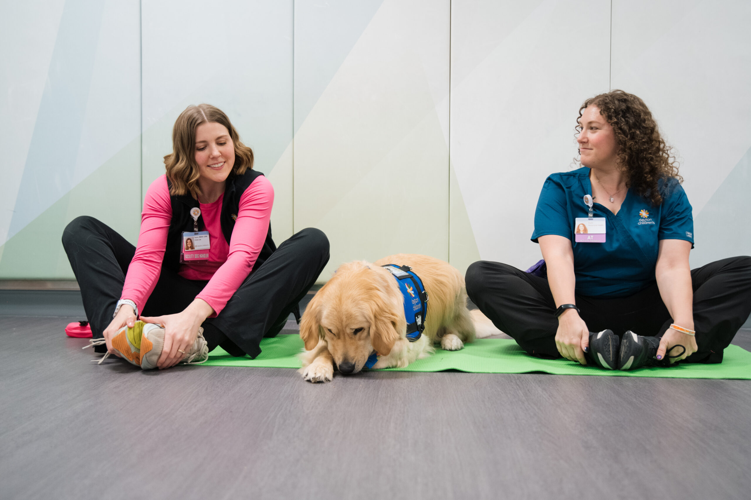A child life specialist and mental health employee sit on a yoga mat with a canine co-pilot doing yoga with a group of patients.. 