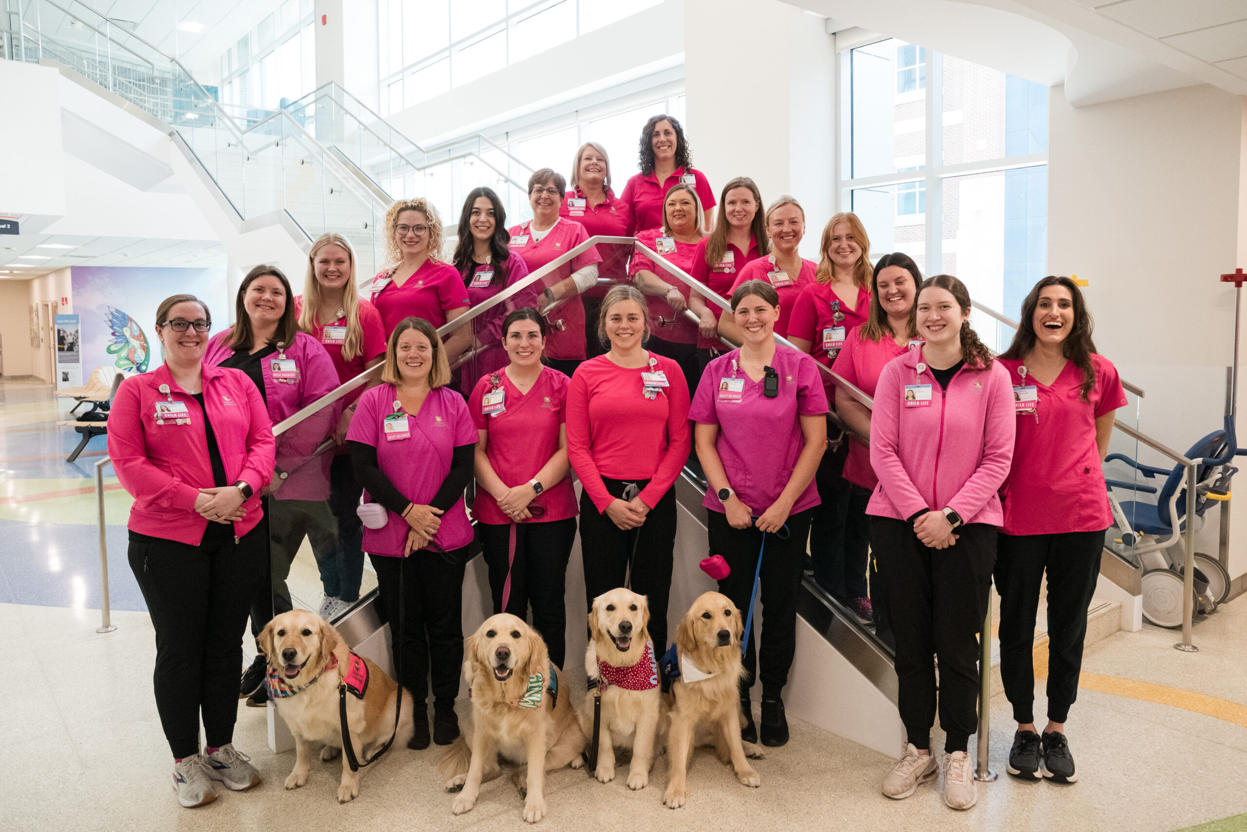 A group of child life employees in their bright pink uniforms  and 4 of the Canine Co-pilot dogs gather for a team photo on the stairs!

