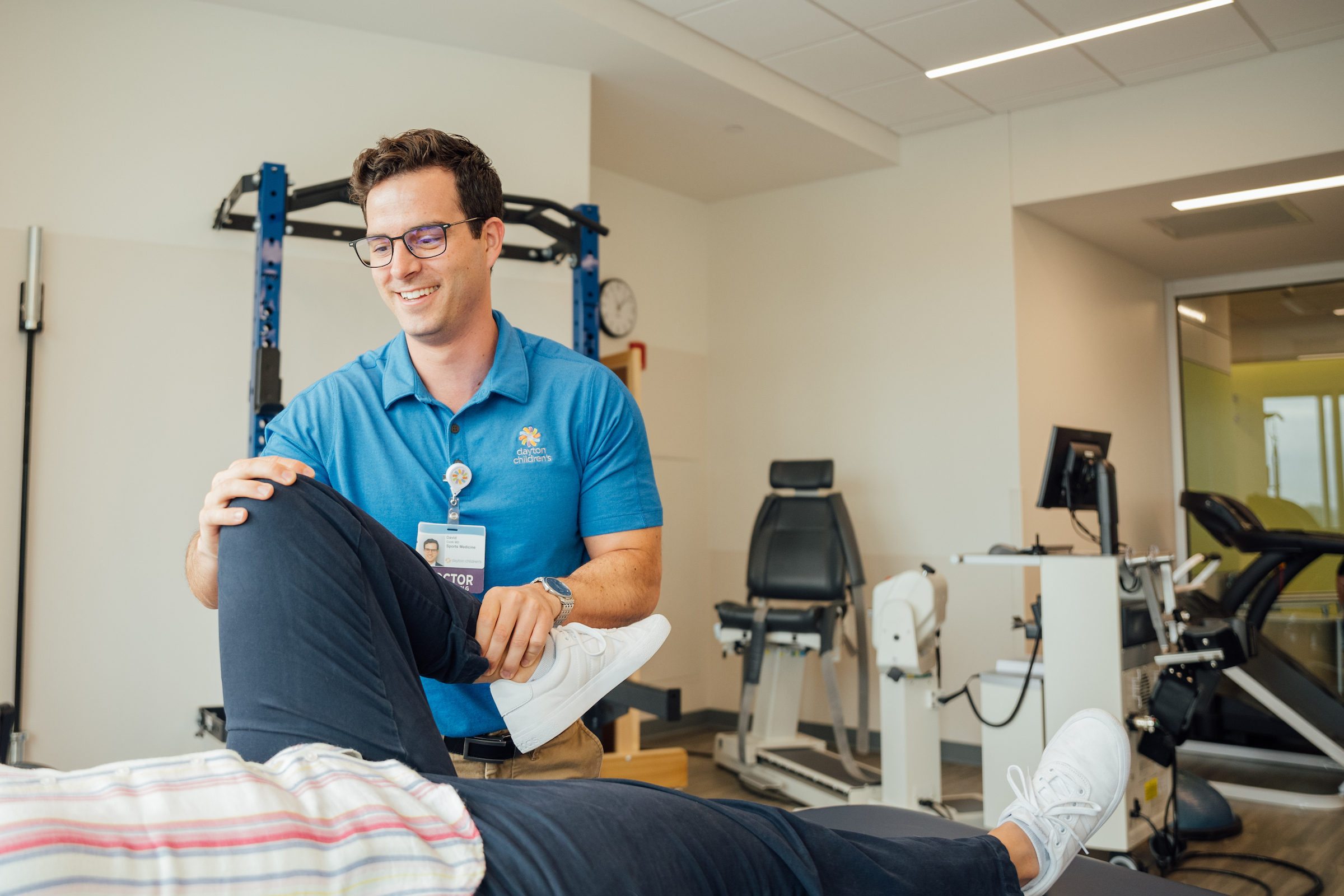 A male doctor wearing a blue Dayton Children's shirt is helping a patient with a knee injury by gently lifting their leg during a physical therapy session.