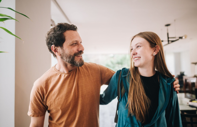 A father and his teenage daughter are smiling happily at each other, with his arm around her shoulder.