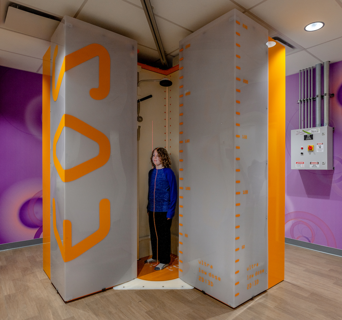 A boy stands between two large columns, facing the camera, with a laser line across his chest for a medical scan.

