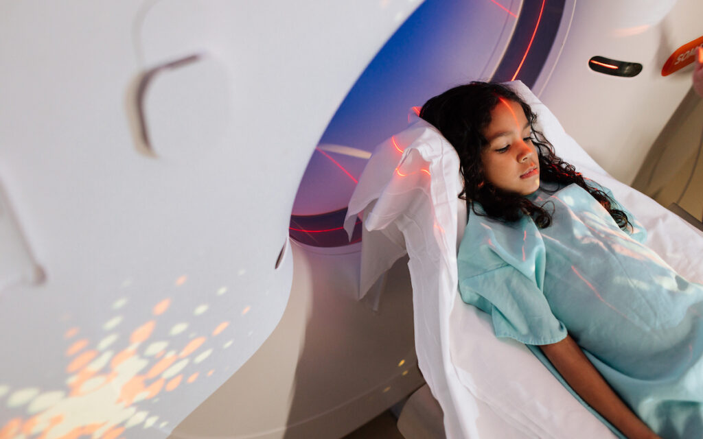 A young girl with long, curly hair lies on a bed, illuminated by red and orange lights from a medical imaging machine.
