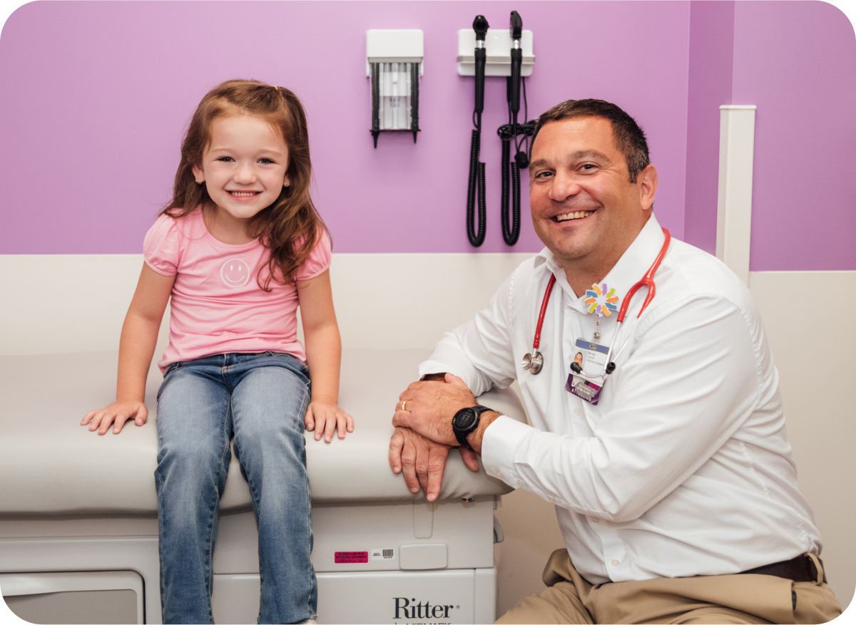 A smiling young girl sits beside a Dayton Children’s provider in a colorful exam room, encouraging families to share their care journey and inspire others.