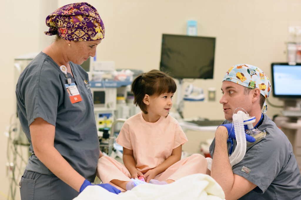 A little girl in a hospital gown on a hospital bed, laughing with two anesthesiologists before surgery.