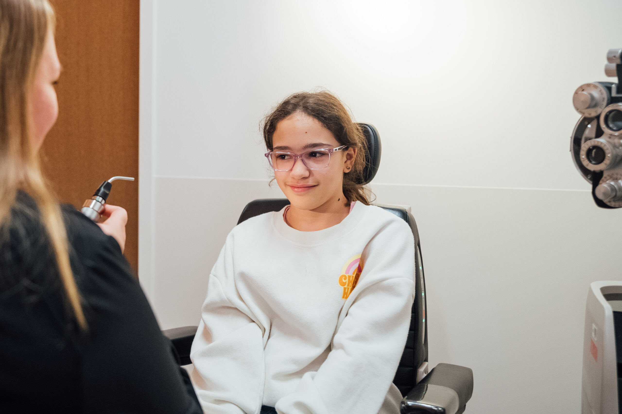 A teen girl sits in the exam chair while the ophthalmologist tests her eye’s reaction to light.