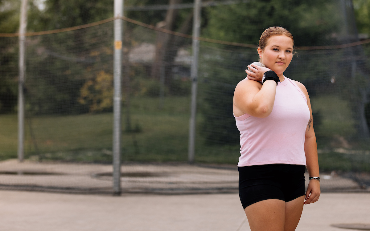 A professional headshot of a female shot athlete in a pink tank top and black shorts, holding a shot put on her shoulder.

