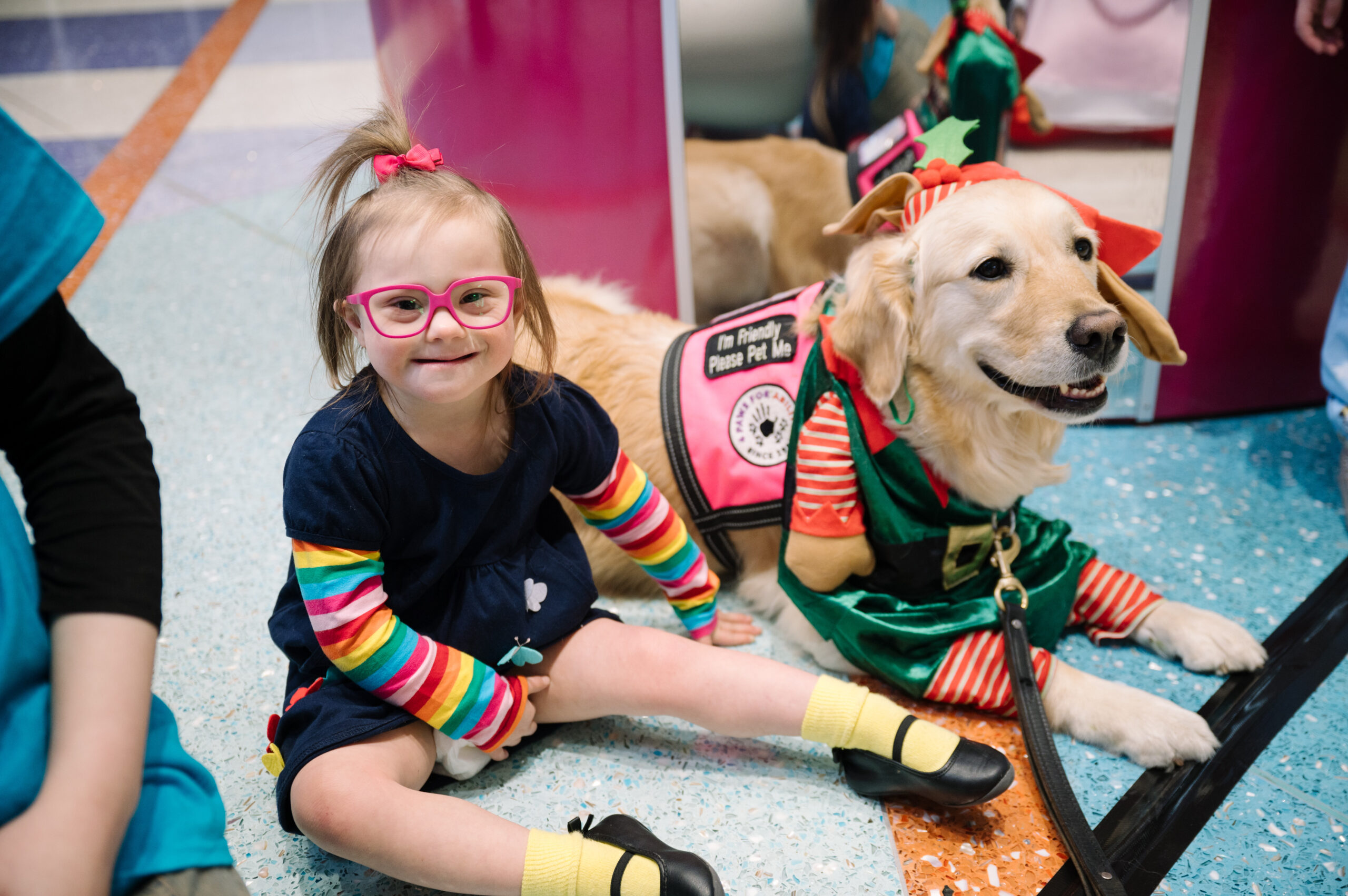 A smiling little girl with down syndrome sitting next to Dayton Children’s Canine Co-pilot, who is dressed as an elf for the holidays.