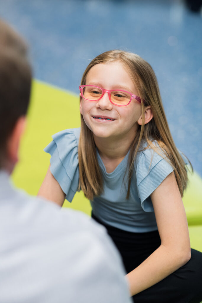 A young girl with long brown hair, wearing pink glasses and a blue ruffled shirt, smiles and looks at someone off-camera.


