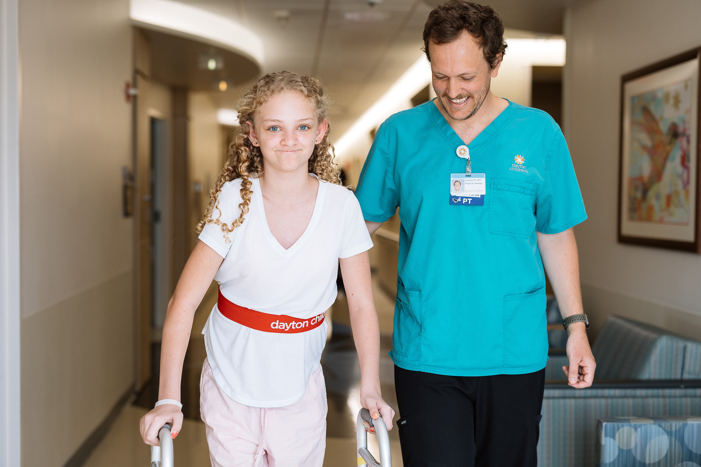 Physical therapist assisting a young patient using a walker during a rehabilitation session in a hospital hallway.