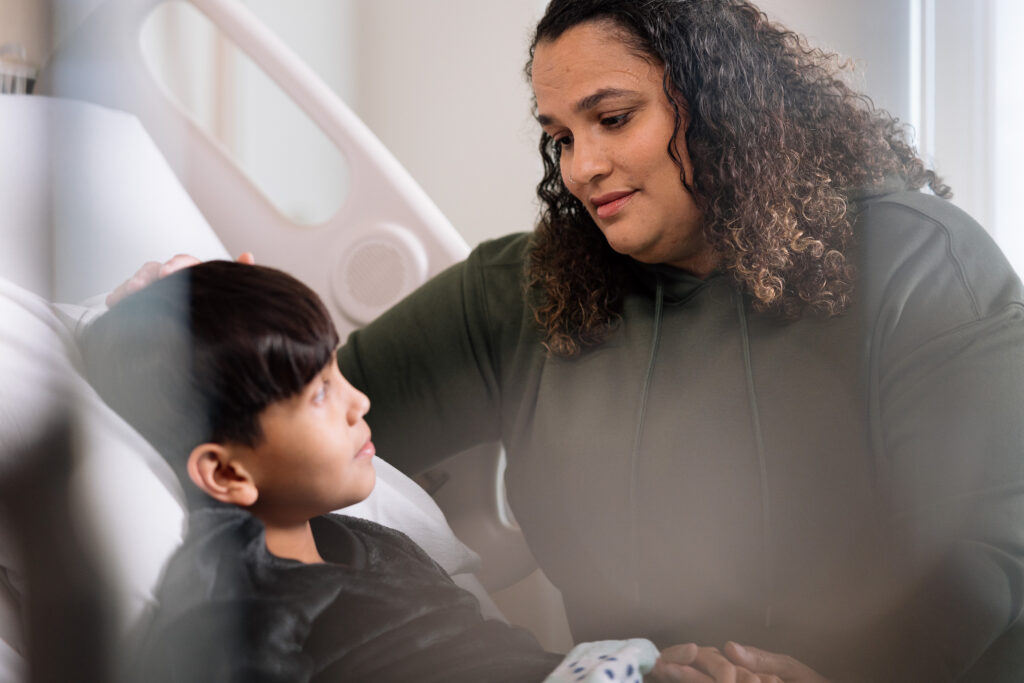 A caring woman with curly hair gently touches the head of a young boy lying in a hospital bed, both looking at each other.

