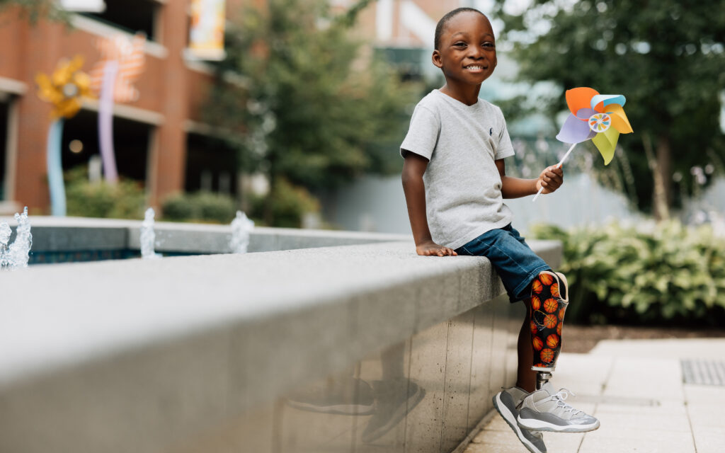 A smiling young boy holds a colorful pinwheel while sitting near a fountain.