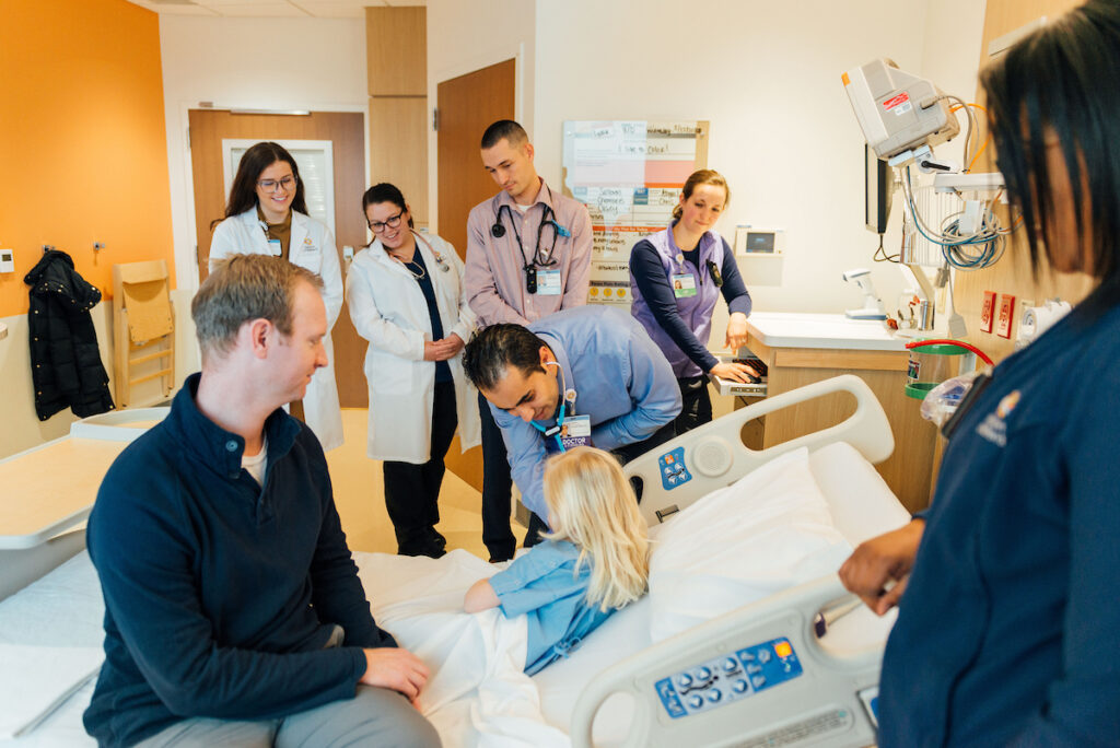A medical team, including several doctors and nurses, attends to a young child in a hospital bed while a man sits nearby.
