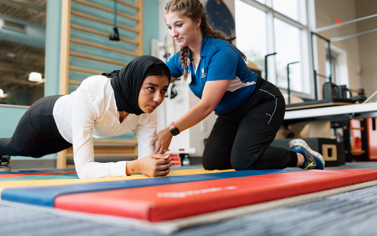 A physical therapist helps a young woman maintain a plank position on a mat.