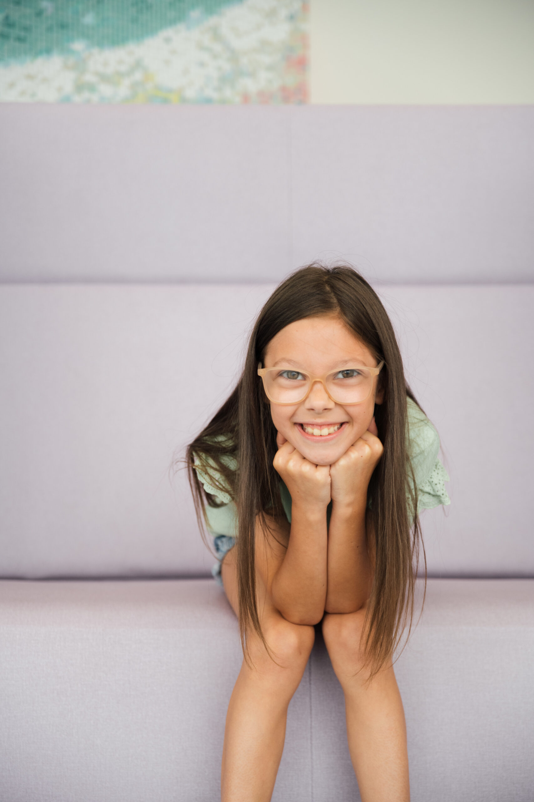 A girl on a purple couch smiling at the camera wearing her glasses, waiting for her appointment. 