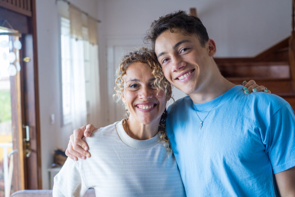 A happy mother with curly hair and her teenage son with a lightning bolt necklace stand together with their arms around each other, smiling at the camera.