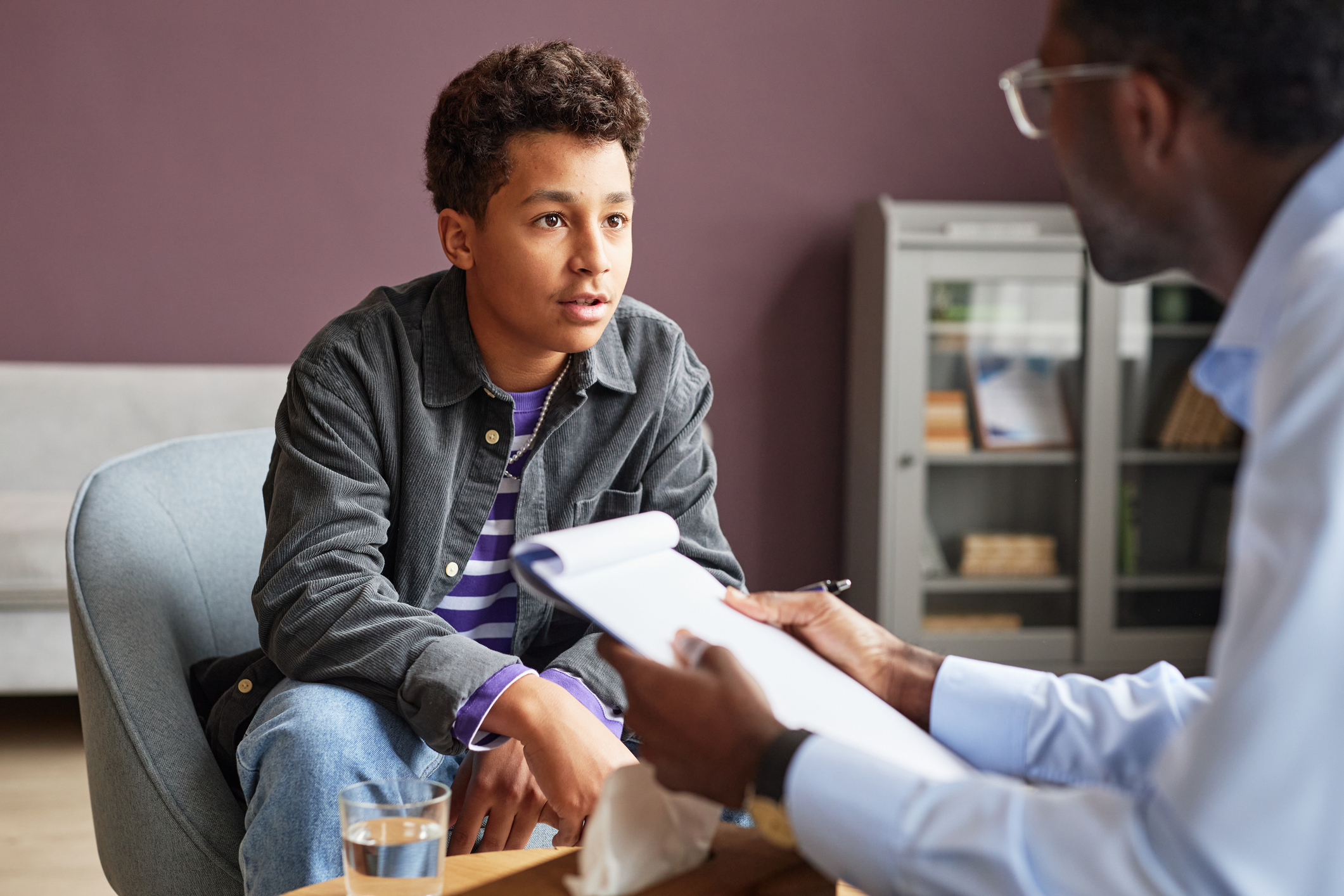 A boy with tourettes is seated in a chair, talking to a medical professional who is holding a clipboard. 