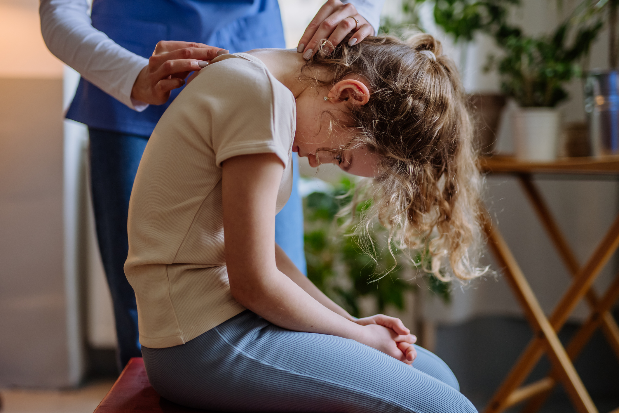 A medical professional in blue scrubs is examining a young girl's spine, as she bends forward. 