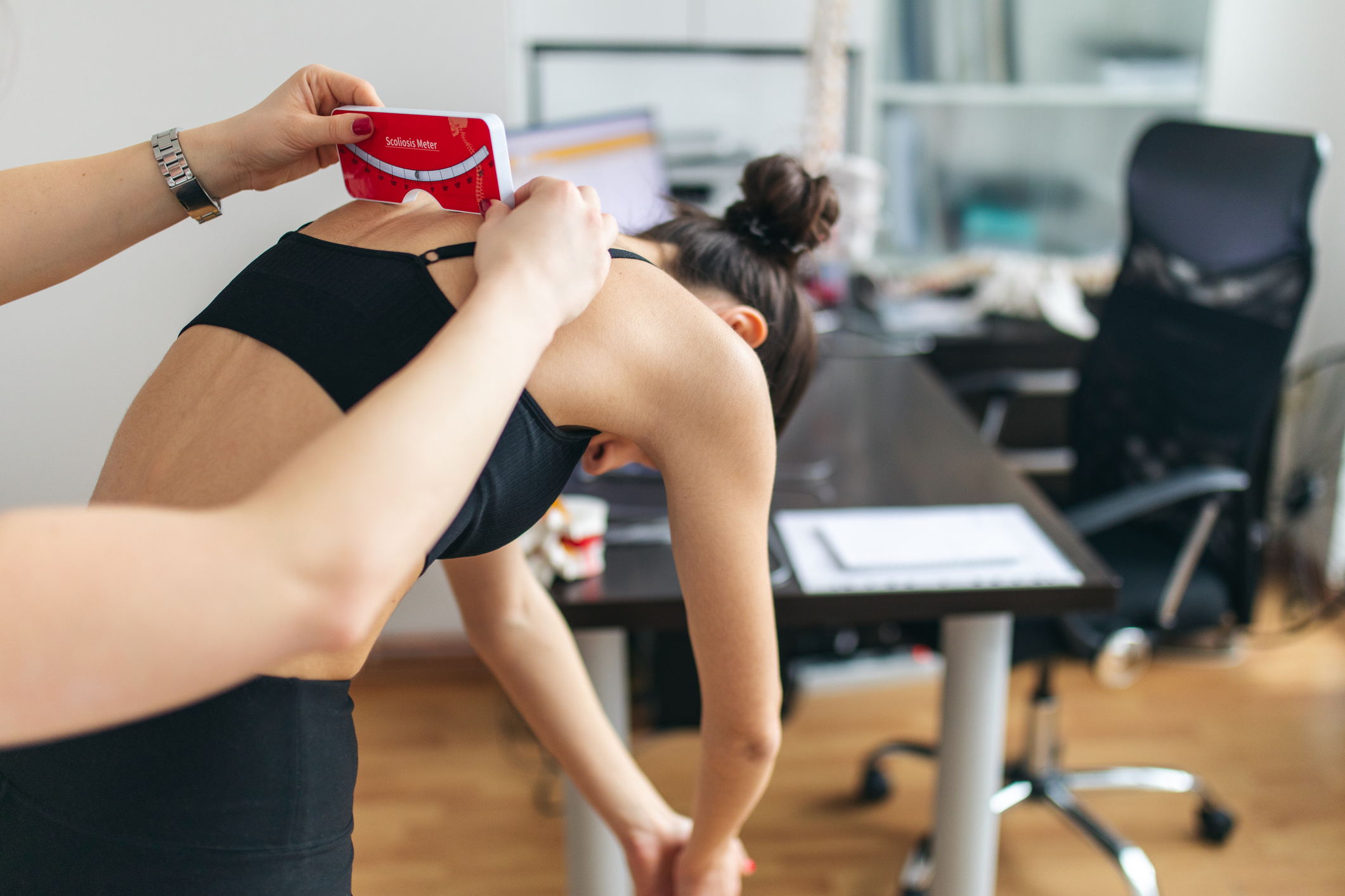 A patient with their back to the camera is being examined for scoliosis using a "Scoliometer" tool.