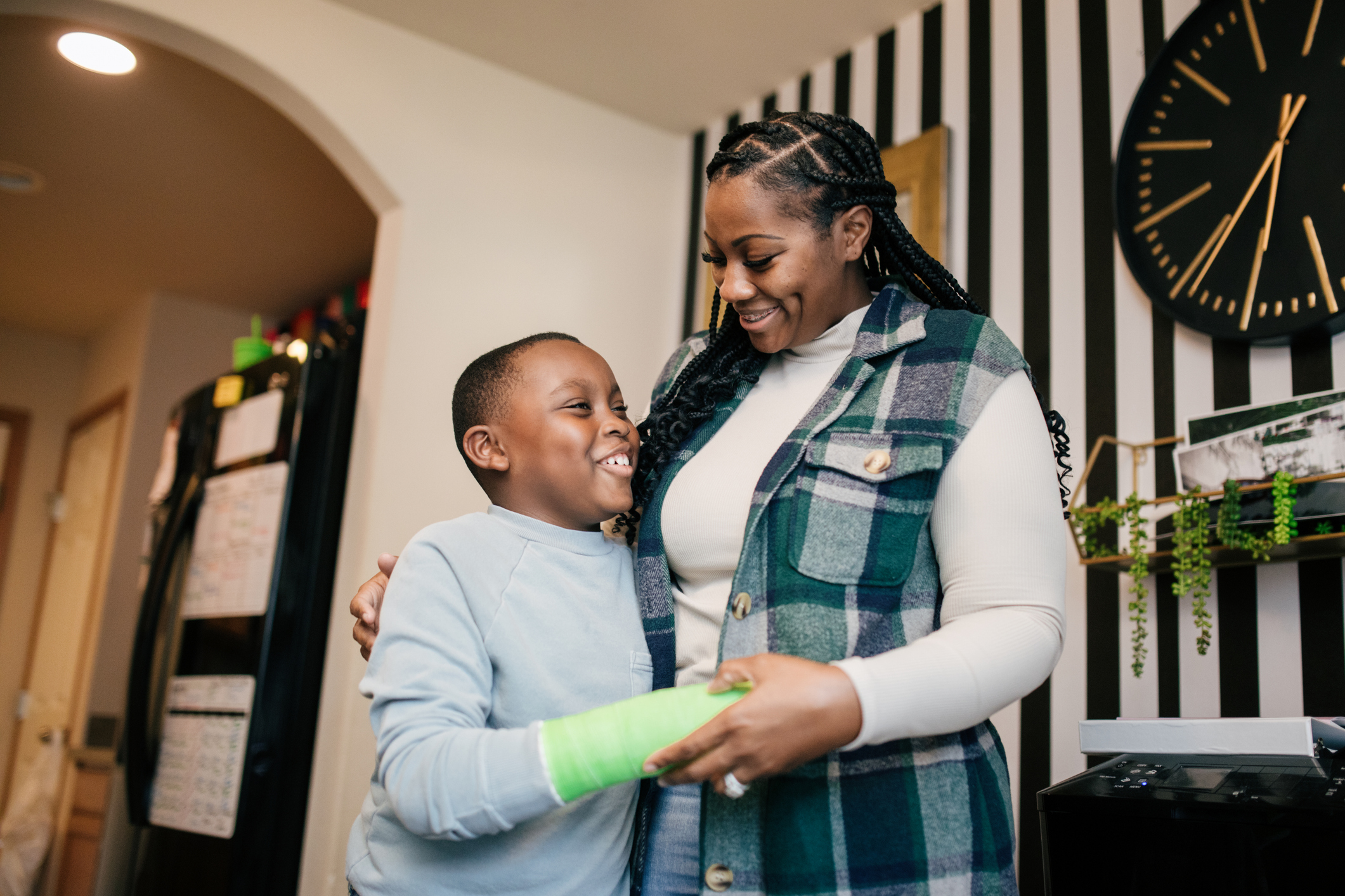 A happy mother and her son are standing together, smiling and embracing. The boy is holding his arm, which is in a bright green cast due to a fracture.