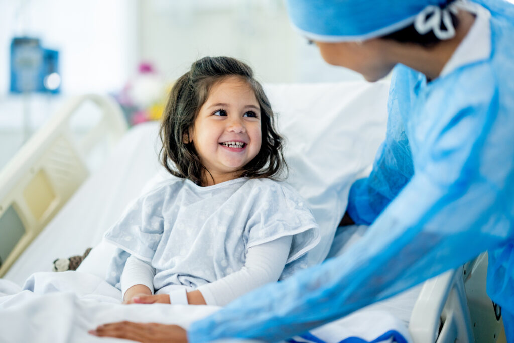 A happy little girl in a hospital gown is sitting up in a hospital bed, smiling at a medical professional who is leaning over her.