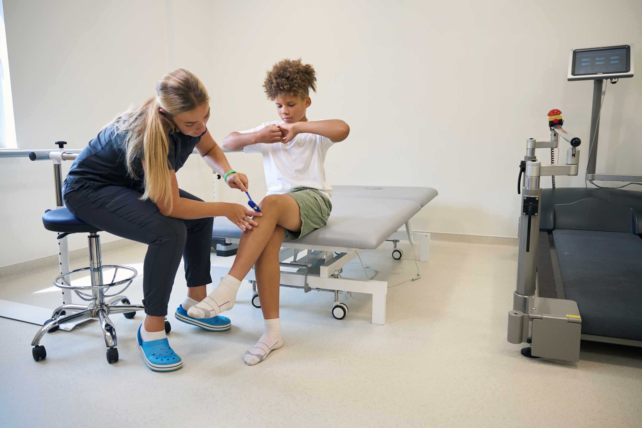 A female physical therapist uses a reflex hammer to examine a boy's knee, likely as part of a physical therapy session for knee injuries.