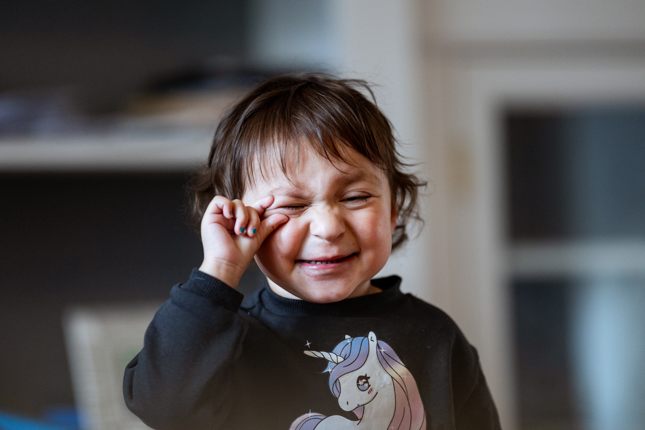 A child in a unicorn sweatshirt is squinting and rubbing their eye with one hand, a common facial tic associated with Tourette's syndrome. 