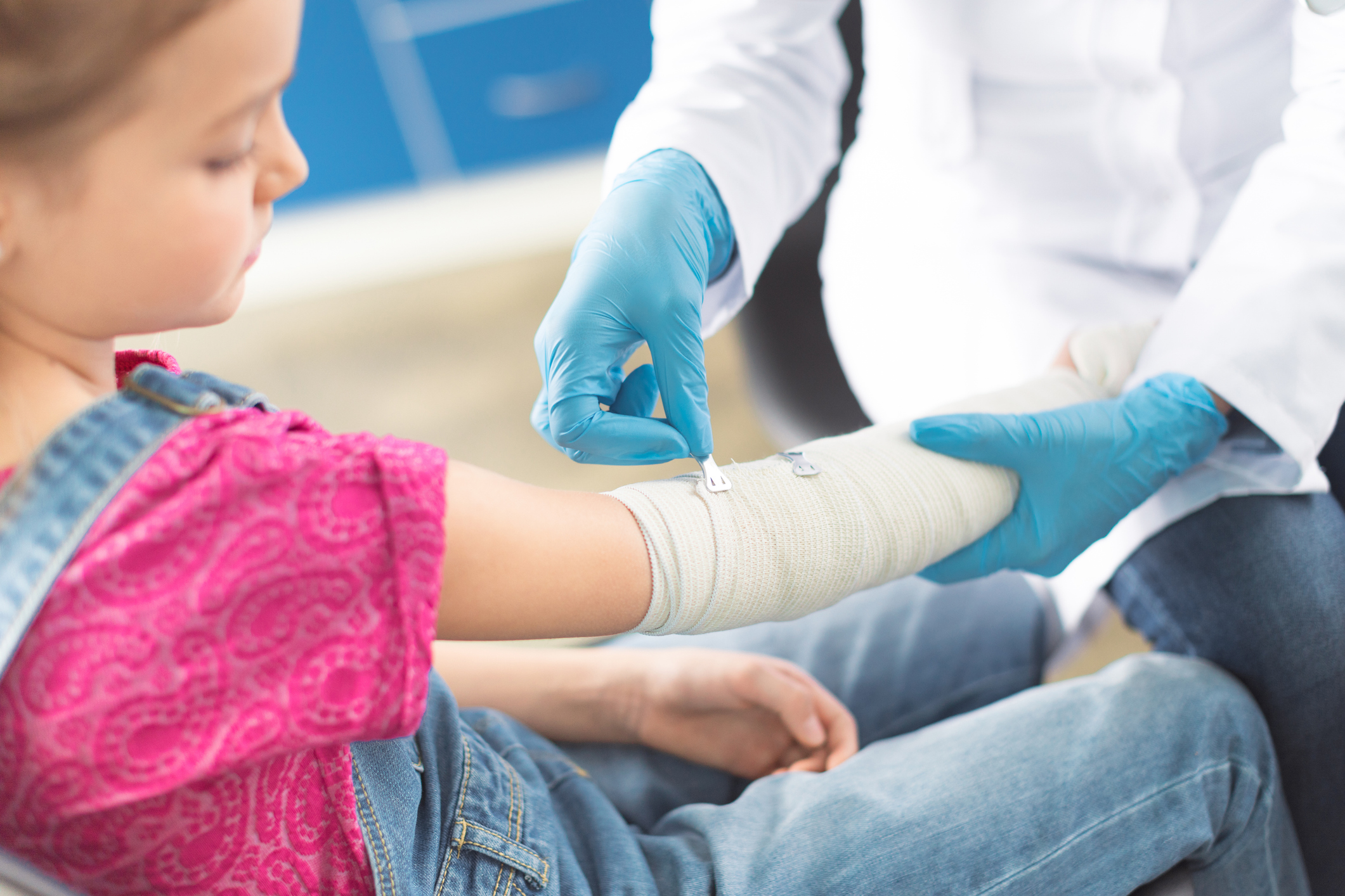 A child with a fracture on their arm has it wrapped in a bandage while a medical professional in blue gloves secures it. 
