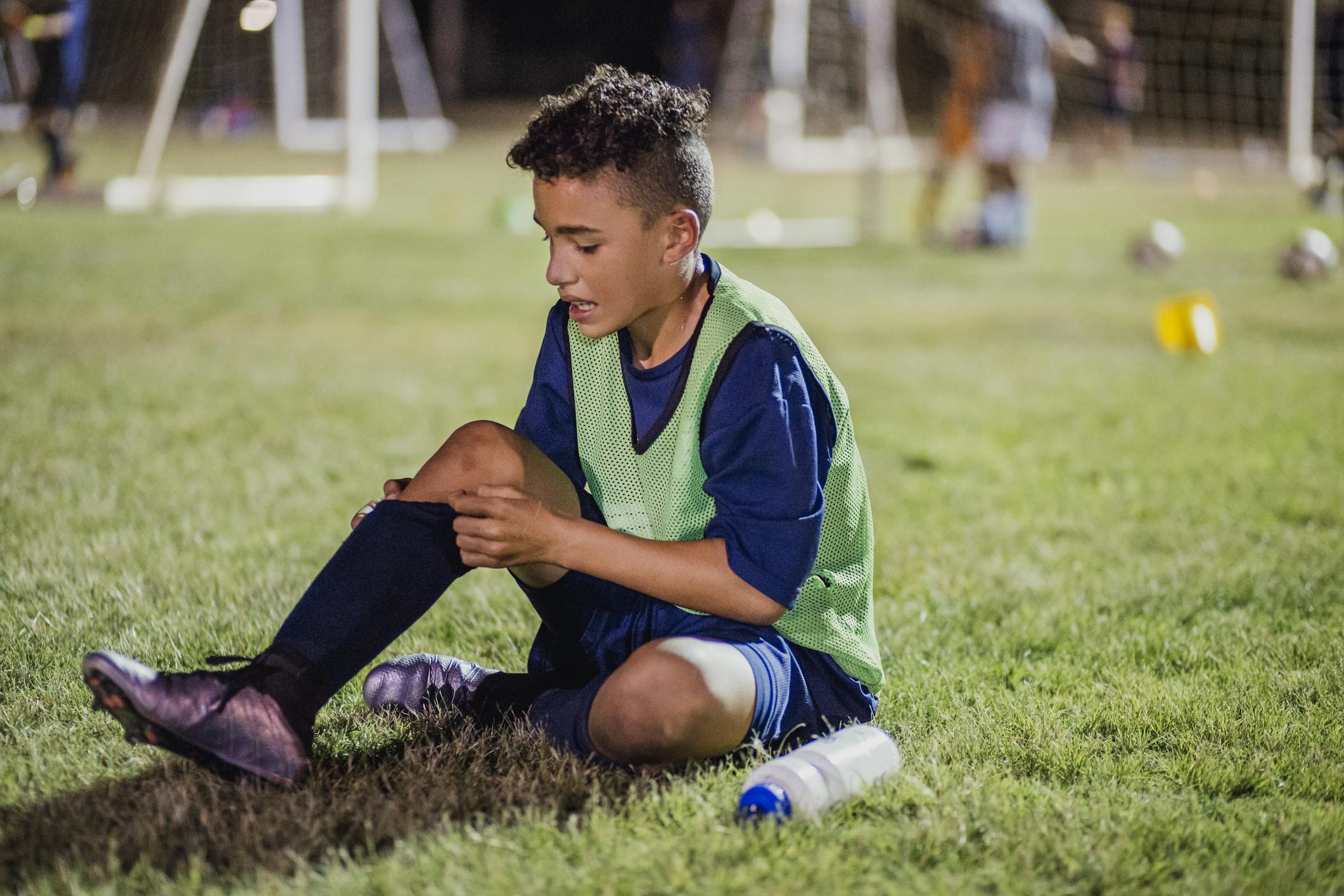 A young male soccer player is sitting on a grassy field, holding his leg with a pained expression.