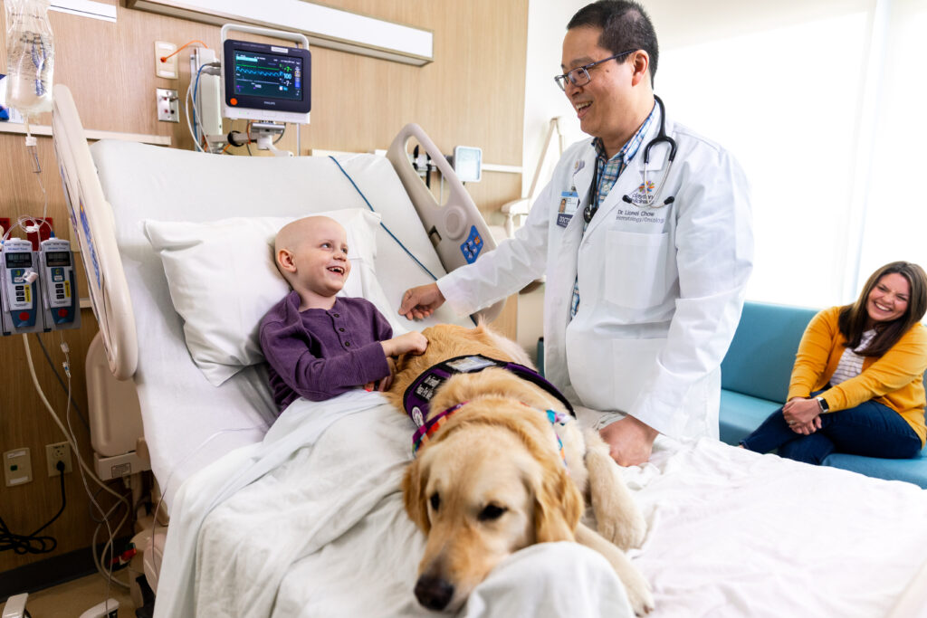A young cancer patient laughs along with the male hematology oncology doctor about the canine copilot therapy dog laying on their lap.