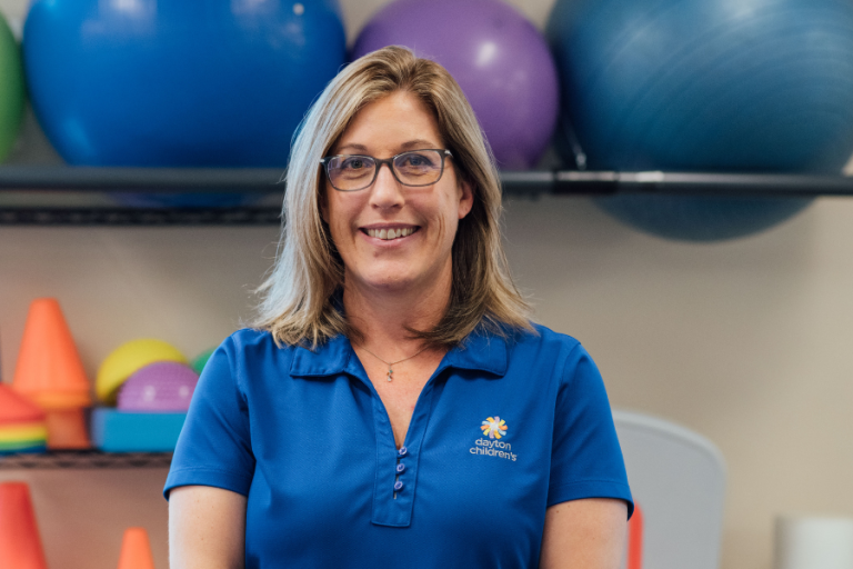 A friendly woman wearing glasses and a blue Dayton Children's polo shirt smiles in a gym setting with large exercise balls behind her.