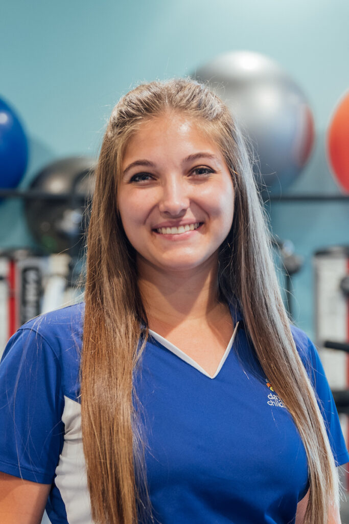 A smiling young woman with long, straight brown hair, wearing a blue and white athletic polo shirt, in a therapy gym setting.