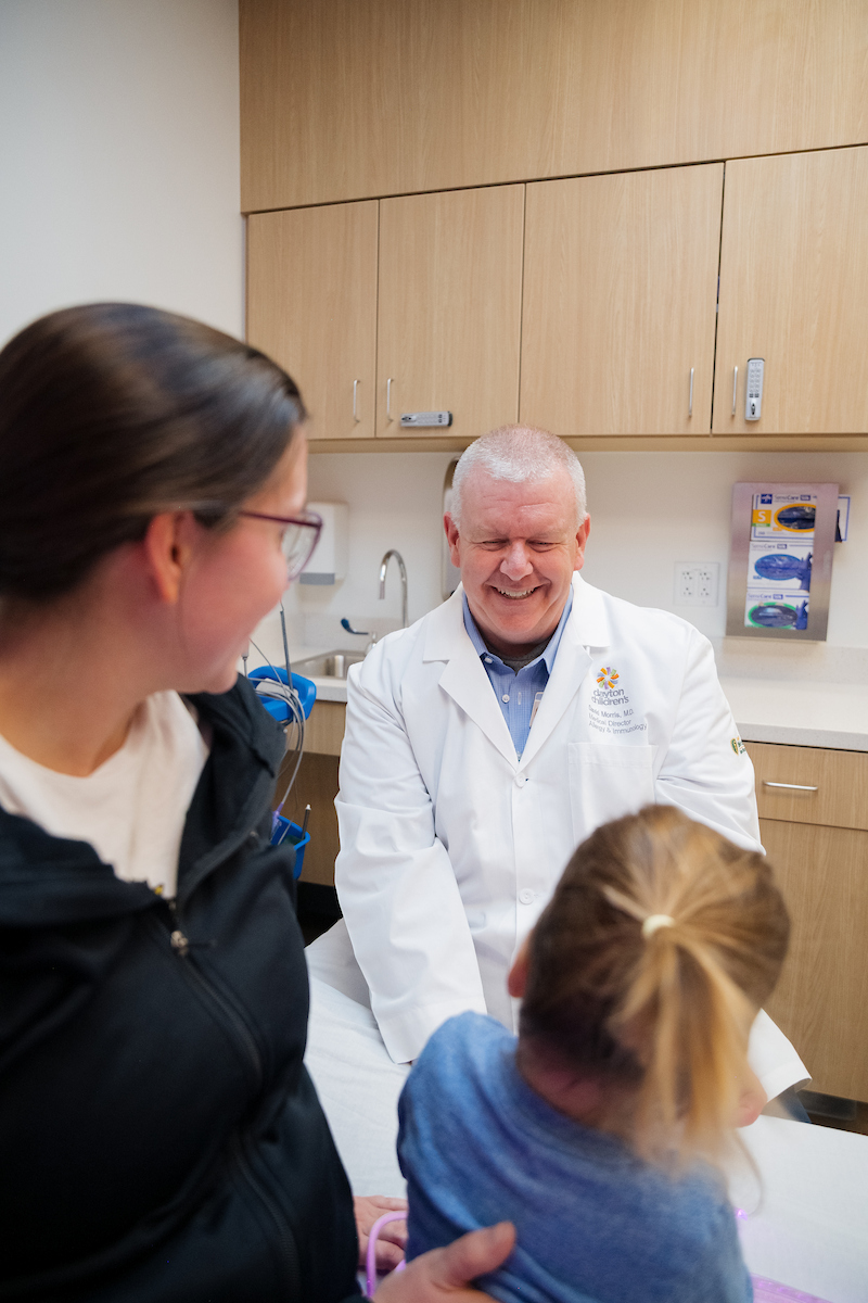 Smiling doctor in a white coat talking with a parent and child in a medical exam room.