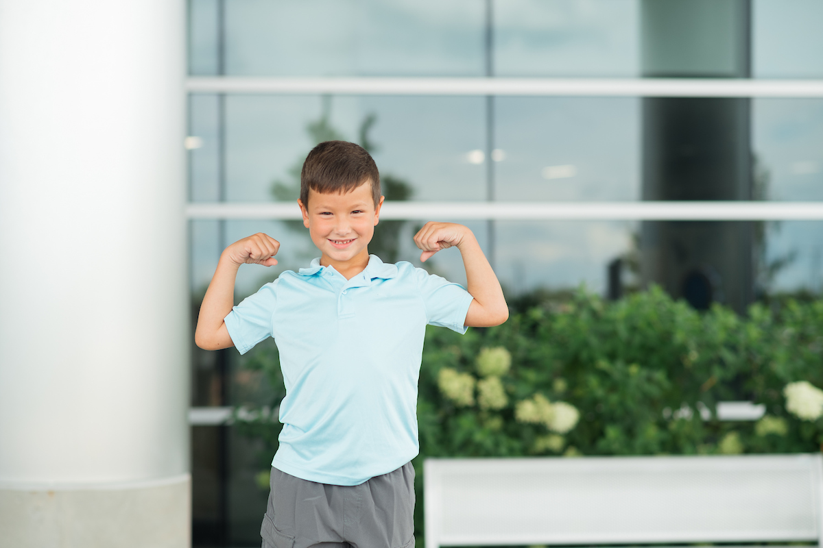 A smiling young boy in a light blue shirt flexing his arms outdoors.

