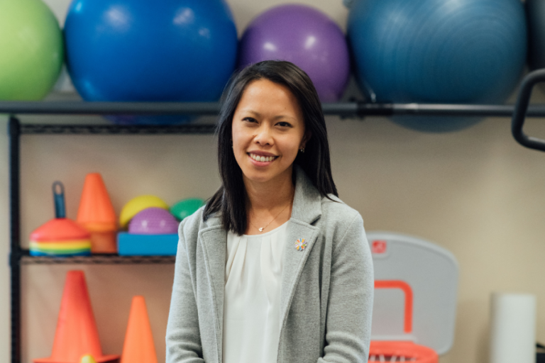 A woman smiles in a gray blazer over a white blouse, wearing a small Dayton Children's pin, in a therapy gym setting.
